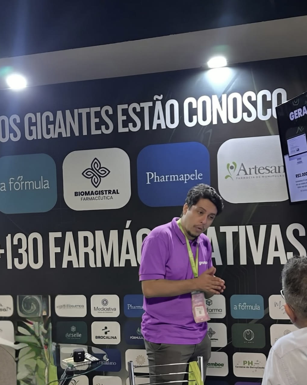 Consultor da Groto no estande da Consulfarma 2026, em frente ao painel com as marcas atendidas
